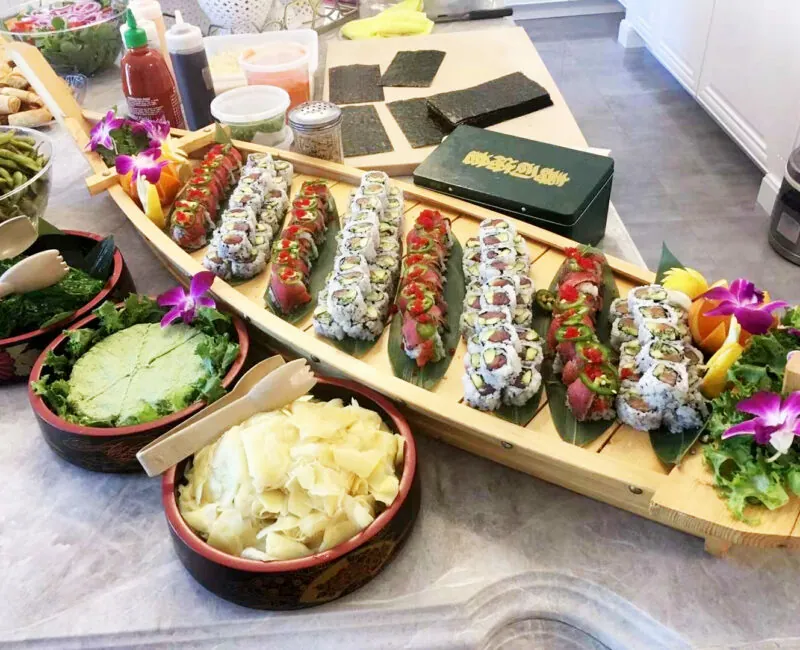 Sushi boat filled with assorted rolls on a counter, orchids and citrus garnish, with bowls of ginger and greens beside it.