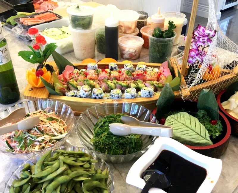 Colorful sushi rolls on a wooden boat, surrounded by edamame, seaweed salad and soy sauce at a catering setup.