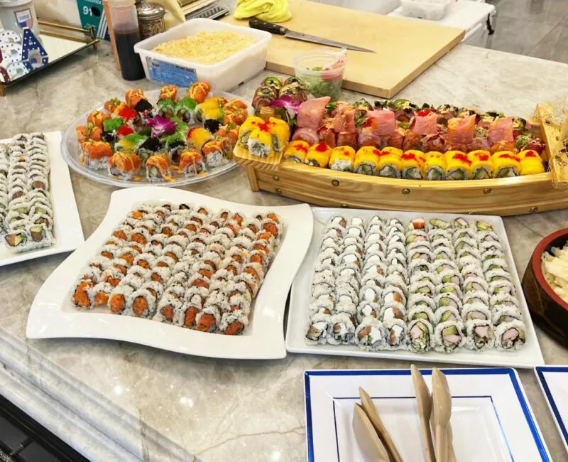 Party trays of neatly arranged sushi rolls on white platters with a wooden sushi boat full of rainbow rolls behind.